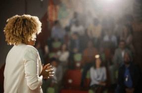 A women facing away from the camera and talking to a large audience. The audience is out of focus.