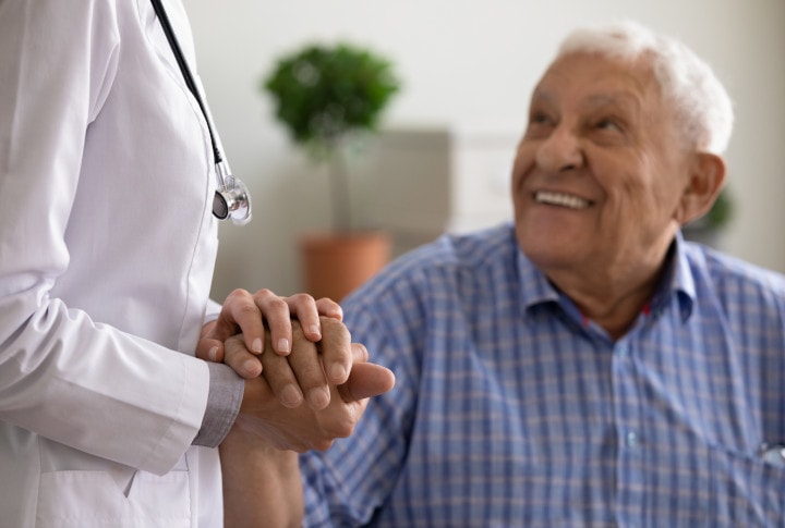 A senior man with a joyful expression looking up at a healthcare professional whose back is to the camera, hands clasped in a comforting gesture.