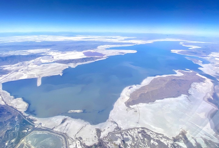 Aerial view of the Great Salt Lake in Utah, showing its vast expanse with varying shades of blue and white salt deposits along the shores, surrounded by a rugged landscape.